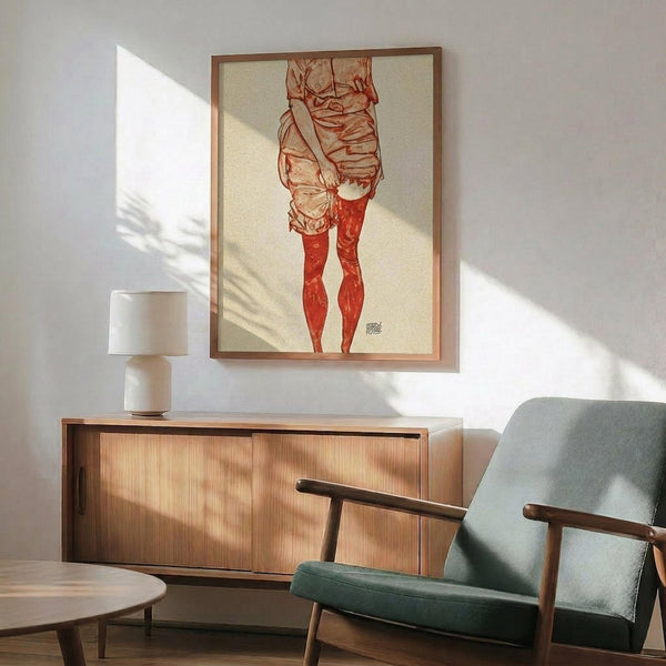 May include: Framed artwork showing a figure in loose clothing with red stockings, displayed above a wooden sideboard with white lamp in modern living room setting with sage green armchair and natural lighting creating wall shadows