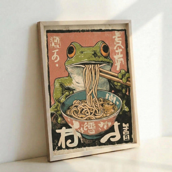 May include: A framed poster print leaning against a white wall on a white surface. The vintage-style artwork features a green frog with large orange eyes eating noodles from a colorful ramen bowl using chopsticks, set against a coral pink background with Japanese characters. The illustration has a weathered, retro aesthetic with muted colors and is displayed in a natural wood frame.