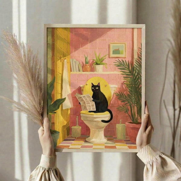 May include: Hands holding a framed illustration showing a black cat sitting on a toilet reading a newspaper in a colorful retro-style bathroom with pink and coral tiled walls, yellow accents, houseplants on a shelf, and checkered floor tiles. The artwork has a mid-century modern aesthetic with warm tones and is displayed in a light wood frame against white curtains, with dried pampas grass visible to the left.