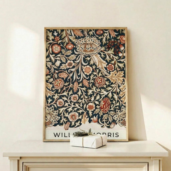 May include: Framed vintage botanical poster featuring William Morris's intricate floral pattern with ornate flowers, leaves, and vines in coral, cream, and gold tones on dark navy background. The print sits on a light wood console table next to a small white wrapped gift with evergreen sprig. The frame appears natural wood against a cream wall.
