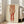 May include: Large framed poster print of an Egon Schiele drawing showing a figure from torso down wearing red stockings and loose clothing, rendered in reddish-brown tones on cream background. White frame leans against wall in bright living room with beige sofa, large windows, and natural lighting creating soft shadows on white walls and light wooden floor.