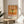 May include: Orange vintage-style poster with green dragonfly illustration displayed in wooden frame on round wooden stool next to beige upholstered chair. Japanese text at top, "DRAGONFLY" and "KYOTO SCIENCE MUSEUM 2005年4月16日" below insect. Sunlight streams across white wall creating shadows from nearby plant leaves.