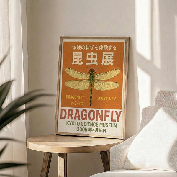 May include: Orange vintage-style poster with green dragonfly illustration displayed in wooden frame on round wooden stool next to beige upholstered chair. Japanese text at top, "DRAGONFLY" and "KYOTO SCIENCE MUSEUM 2005年4月16日" below insect. Sunlight streams across white wall creating shadows from nearby plant leaves.