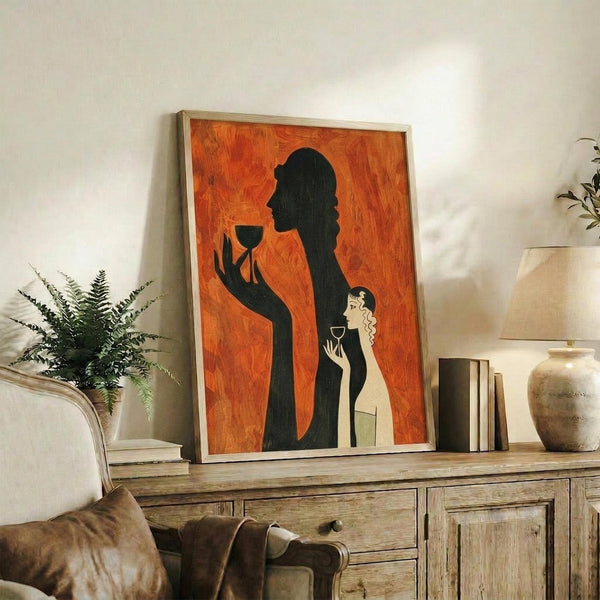 May include: Framed art print with orange background showing black silhouette of woman holding wine glass and smaller light-colored figure, displayed on rustic wooden sideboard alongside fern plant, books, ceramic vase and lamp in modern living room setting with beige armchair