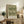 May include: Framed botanical print with intricate green and cream acanthus leaf pattern displayed on rustic wooden sideboard alongside fern plant, stacked books, and ceramic vases in warmly lit living room with beige upholstered furniture and neutral walls