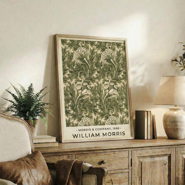 May include: Framed botanical print with intricate green and cream acanthus leaf pattern displayed on rustic wooden sideboard alongside fern plant, stacked books, and ceramic vases in warmly lit living room with beige upholstered furniture and neutral walls