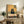 May include: Framed artwork displayed on rustic wooden sideboard showing black cat with glowing yellow eyes sitting at checkered tablecloth-covered table with pizza slices, rendered in warm golden and muted green tones. Scene includes fern plant, ceramic vase, books, and cream-colored armchair in cozy living room setting with white walls.