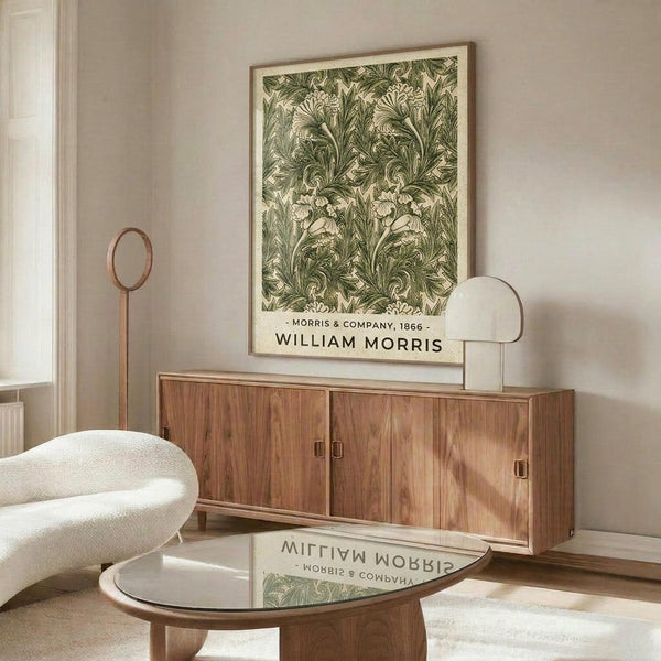 May include: Framed botanical print with green acanthus leaf pattern displayed above a wooden sideboard in modern living room. Print features intricate Victorian-style foliage design in sage green and cream tones. Room styled with curved white chair, round glass coffee table, and brass accent lighting on neutral walls.