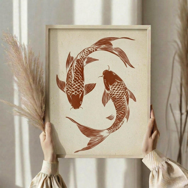 May include: Person holding framed wall art featuring two brown koi fish arranged in circular yin yang pattern on cream textured background, displayed in light wood frame. One hand also holds dried pampas grass stems. Soft neutral room setting with white curtains and beige clothing visible.