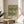 May include: Framed botanical print featuring intricate green foliage pattern with cream background, displayed on round wooden side table next to beige upholstered chair. Natural sunlight creates soft shadows on neutral wall. Plant leaves visible in corner, creating warm minimalist home decor setting.