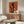 May include: Orange and black Art Deco style poster showing silhouette of woman holding wine glass with smaller blonde woman figure below, displayed above wood credenza in modern living room with white curved chair, oval glass coffee table, and white table lamp