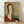 May include: Framed reproduction of a 1917 portrait painting showing a woman with an elongated face and neck in Modigliani's distinctive style. She has dark bobbed hair, wears a deep red V-neck garment, and sits against a muted olive-green background. The artwork is displayed in a light wooden frame, leaning against a white wall on light wood flooring with natural shadows visible.