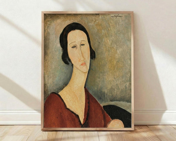 May include: Framed reproduction of a 1917 portrait painting showing a woman with an elongated face and neck in Modigliani's distinctive style. She has dark bobbed hair, wears a deep red V-neck garment, and sits against a muted olive-green background. The artwork is displayed in a light wooden frame, leaning against a white wall on light wood flooring with natural shadows visible.