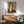 May include: Framed portrait print hanging on white wall above wooden sideboard in modern living room setting. Artwork shows woman with elongated face and neck, dark hair, wearing burgundy top against muted background in characteristic early 20th century style. Mid-century furniture includes teal upholstered armchair with wooden frame, walnut credenza, ceramic table lamp, and round coffee table with natural sunlight streaming across scene.