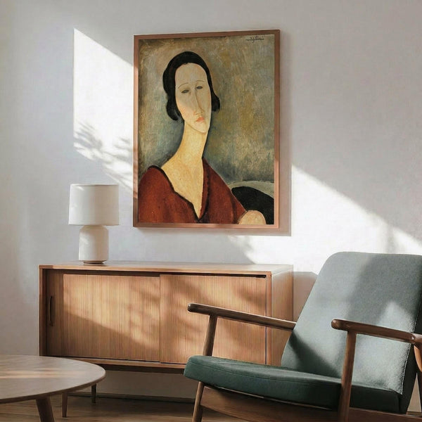 May include: Framed portrait print hanging on white wall above wooden sideboard in modern living room setting. Artwork shows woman with elongated face and neck, dark hair, wearing burgundy top against muted background in characteristic early 20th century style. Mid-century furniture includes teal upholstered armchair with wooden frame, walnut credenza, ceramic table lamp, and round coffee table with natural sunlight streaming across scene.
