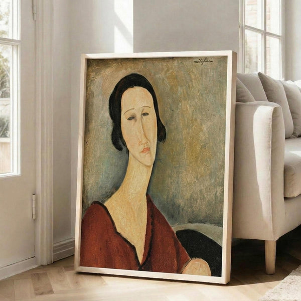 May include: Framed art print featuring a portrait of a woman with black hair and elongated neck wearing a deep red garment, painted in modernist style with muted earth tones and sage green background. Large rectangular frame leans against white wall next to beige upholstered chair in bright room with tall windows.