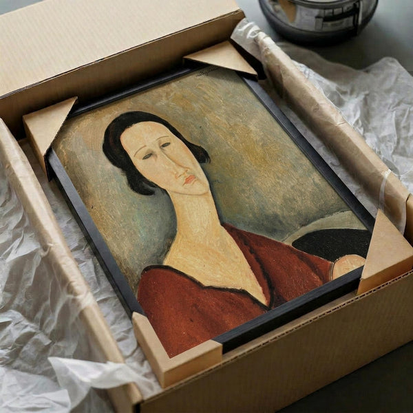 Framed art print of a woman with an elongated neck and face in Modigliani's distinctive style, featuring muted earth tones of rust red, olive green, and cream. The portrait shows a serene female figure with dark hair and closed eyes against a soft background. The print is being carefully unpacked from a cardboard shipping box with protective tissue paper and wooden corner guards visible around the black-framed artwork.