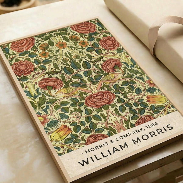 Vintage botanical poster featuring an intricate floral pattern with coral pink roses, green leaves, and flowing vines on a cream background. The decorative design shows William Morris & Company 1866 text at the bottom. The print sits on white paper with a cream ribbon beside it, displaying the classic Arts and Crafts movement style with detailed hand-drawn elements and rich earth tones.