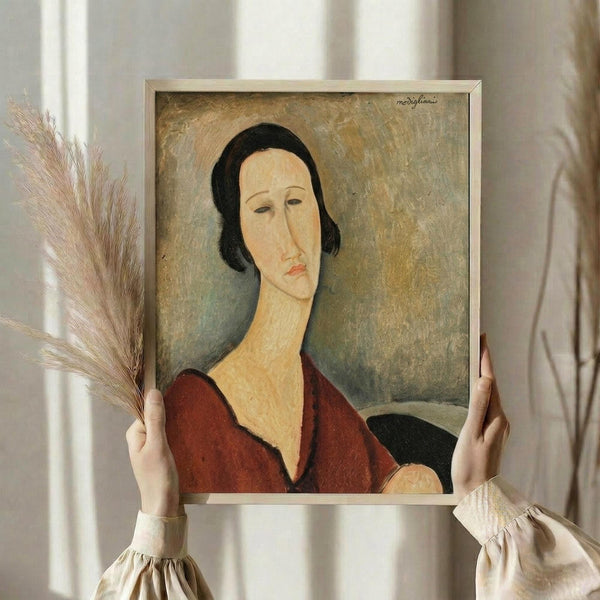 May include: Hands holding a framed reproduction of a Modigliani portrait showing a woman with an elongated face, dark bob haircut, and red dress against a muted background. The thin silver frame is displayed against white curtains with dried wheat stems visible on the left side, creating a soft, natural styling scene.