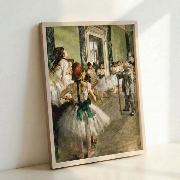 May include: Framed reproduction of Degas ballet painting showing young dancers in white tutus with green sashes practicing in an elegant room with pale green walls, dark curtains, and wooden floors. An instructor observes from the right while ballerinas stretch and pose. The print is displayed in a light wood frame leaning against a white wall.