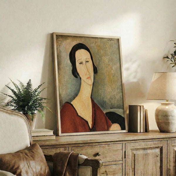 May include: Framed portrait print displayed on rustic wooden sideboard in neutral living room. Artwork shows woman with elongated face, black hair, and burgundy V-neck top in Modigliani's distinctive style with muted earth tones. Scene includes cream upholstered chair, fern plant, ceramic vase, stacked books, and soft natural lighting creating warm shadows on white walls.