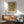 May include: Framed vintage botanical poster with red roses and green foliage pattern hanging above wooden sideboard, mid-century modern sage green armchair with wooden arms, white ceramic table lamp, and round coffee table in bright sunlit room with white walls