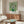 May include: Van Gogh roses poster print in thin wooden frame hanging above mid-century walnut credenza in modern living room. White roses in green vase against lime green background. Room features cream upholstered swivel chair, brass floor lamp, oval glass coffee table with colorful abstract art surface, white walls, and natural lighting from window.