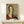 May include: Framed reproduction of a 1917 portrait painting showing a woman with an elongated face and neck in Modigliani's distinctive style, wearing a deep red garment against a muted gray-green background. The artwork is displayed in a thin wooden frame on a light wood surface alongside a small white wrapped gift with evergreen sprig, set against cream-colored walls with soft natural lighting.