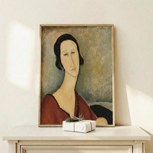 May include: Framed reproduction of a 1917 portrait painting showing a woman with an elongated face and neck in Modigliani's distinctive style, wearing a deep red garment against a muted gray-green background. The artwork is displayed in a thin wooden frame on a light wood surface alongside a small white wrapped gift with evergreen sprig, set against cream-colored walls with soft natural lighting.
