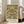 May include: Framed vintage botanical poster with ornate floral pattern featuring deep red roses, green leaves, and golden yellow accents in symmetrical Arts and Crafts style design. The artwork is displayed in a light wood frame, leaning against white walls near large windows in a bright living room with beige upholstered armchair and wooden floors.