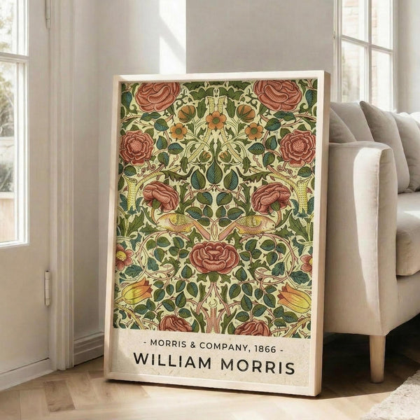 May include: Framed vintage botanical poster with ornate floral pattern featuring deep red roses, green leaves, and golden yellow accents in symmetrical Arts and Crafts style design. The artwork is displayed in a light wood frame, leaning against white walls near large windows in a bright living room with beige upholstered armchair and wooden floors.