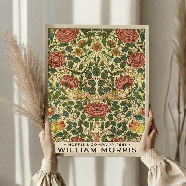 May include: Person holding framed poster featuring William Morris floral pattern with red roses, green leaves, and cream background in Arts and Crafts style. Light wood frame against soft neutral curtained background with dried pampas grass visible on left side. Vintage botanical wallpaper design from Morris & Company 1866.