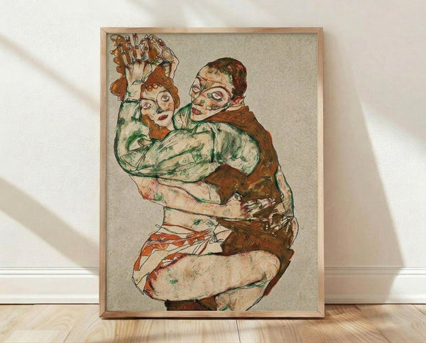 May include: Framed reproduction of an expressionist artwork showing two embracing figures in muted earth tones of green, brown, and orange against a beige background. The wooden frame sits on light hardwood flooring against white walls with natural sunlight casting diagonal shadows across the scene.