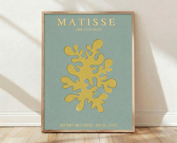 May include: Framed art print with sage green background featuring yellow organic cutout shape resembling coral or seaweed. Text reads "MATISSE THE CUT-OUTS" at top and "HENRI MATISSE, NICE, 1947" at bottom in golden lettering. Natural wood frame sits on light wooden floor against white textured wall with soft shadow patterns from window light.