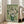 May include: Framed reproduction of Van Gogh's white roses painting displayed in bright living room. Green vase holds abundant white blooms with thick brushstrokes against sage green background. Light wood frame leans against white wall near beige armchair and large windows with natural light streaming across hardwood floors.