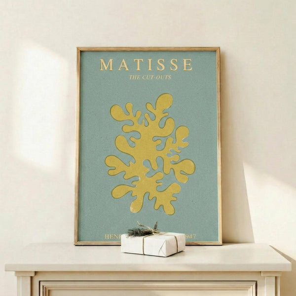 May include: Framed Matisse poster with yellow coral-like abstract cut-out shape on sage green background, displayed on light wood console table with small white wrapped gift box beside it, against cream wall with natural lighting