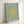 A framed art print featuring a yellow coral-like organic shape on a sage green background. The print displays "MATISSE" and "THE CUT-OUTS" at the top in gold lettering, with "HENRI MATISSE, NICE, 1947" at the bottom. The artwork is mounted in a light wooden frame and photographed leaning against a white wall, showing the frame's depth and natural wood grain texture.