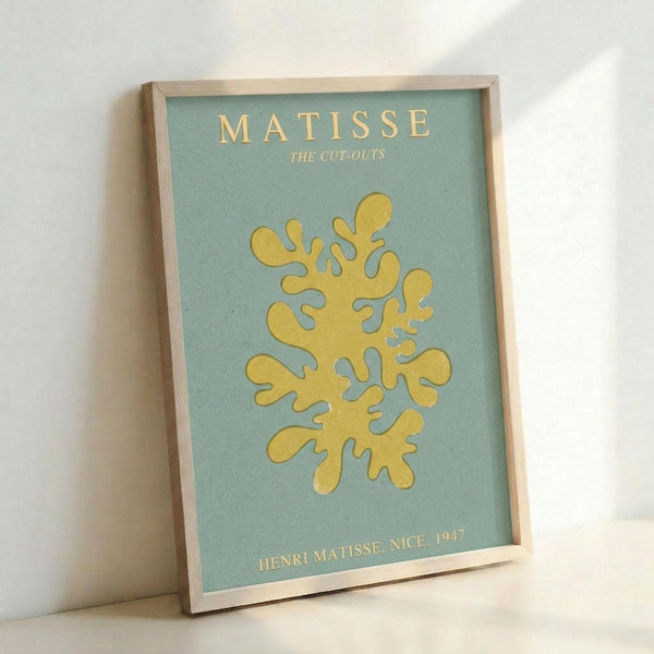 A framed art print featuring a yellow coral-like organic shape on a sage green background. The print displays "MATISSE" and "THE CUT-OUTS" at the top in gold lettering, with "HENRI MATISSE, NICE, 1947" at the bottom. The artwork is mounted in a light wooden frame and photographed leaning against a white wall, showing the frame's depth and natural wood grain texture.