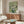 May include: Van Gogh floral painting reproduction featuring white roses in green vase against soft green background, displayed above mid-century modern walnut credenza in minimalist living room with cream chair, oval glass coffee table, and brass floor lamp on light wooden floors.