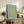 May include: Framed art print with light blue background and yellow organic coral-like cutout shape, displayed on rustic wooden sideboard next to fern plant, ceramic vases, and books in neutral-toned living room setting with beige armchair visible