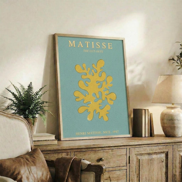 May include: Framed art print with light blue background and yellow organic coral-like cutout shape, displayed on rustic wooden sideboard next to fern plant, ceramic vases, and books in neutral-toned living room setting with beige armchair visible