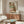 May include: Framed reproduction of early 20th century expressionist artwork showing two embracing figures in earth tones and greens, displayed above a mid-century modern walnut credenza. Room features cream walls, white upholstered chair, glass coffee table, and warm natural lighting creating shadows across the wooden furniture.