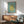 May include: Framed Matisse cut-outs poster with turquoise background and yellow organic coral-like shape, displayed above mid-century wooden sideboard with white ceramic lamp. Gray upholstered armchair with wooden frame sits nearby on hardwood floors. Natural sunlight casts geometric shadows across white walls in modern living room setting.