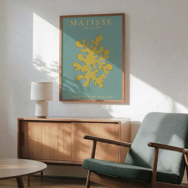 May include: Framed Matisse cut-outs poster with turquoise background and yellow organic coral-like shape, displayed above mid-century wooden sideboard with white ceramic lamp. Gray upholstered armchair with wooden frame sits nearby on hardwood floors. Natural sunlight casts geometric shadows across white walls in modern living room setting.