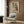 May include: Framed poster print featuring two embracing figures in earth tones of green, brown, and rust, displayed on wood-paneled wall above cream upholstered armchair with geometric pillow. Modern floor lamp with brass finish and round marble base sits nearby alongside dark side table with open book. Warm neutral interior setting with natural lighting.