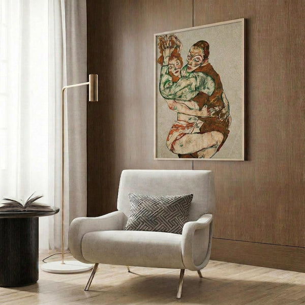May include: Framed poster print featuring two embracing figures in earth tones of green, brown, and rust, displayed on wood-paneled wall above cream upholstered armchair with geometric pillow. Modern floor lamp with brass finish and round marble base sits nearby alongside dark side table with open book. Warm neutral interior setting with natural lighting.