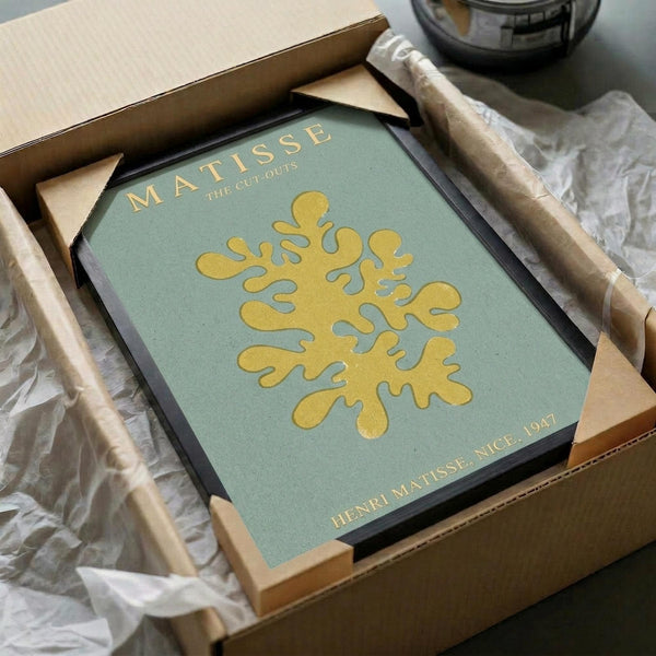May include: Framed Matisse cut-out print featuring a golden yellow organic coral-like shape on sage green background, packaged in brown cardboard box with protective tissue paper. The artwork shows "MATISSE" and "THE CUT-OUTS" text at top, "HENRI MATISSE, NICE, 1947" at bottom. Black frame sits nestled in shipping materials on white surface.