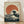 May include: Framed poster print leaning against white wall on light wood floor. Features Japanese woodblock style artwork with large dark blue ocean wave with white foam, orange-red sun in background, and Mount Fuji silhouette. Traditional beige textured paper appearance with Japanese characters in upper right corner. Natural wood frame, soft lighting creates diagonal shadows across scene.