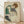 May include: Framed reproduction of an early 20th century expressionist drawing showing a seated woman with curly reddish hair wearing a green sleeveless top and dark stockings. The artwork features loose, sketchy linework on cream-colored paper with an artist's signature stamp. The thin wooden frame sits on light wood flooring against a white wall with soft natural lighting creating diagonal shadows.