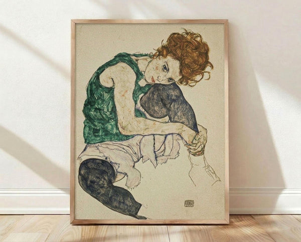 May include: Framed reproduction of an early 20th century expressionist drawing showing a seated woman with curly reddish hair wearing a green sleeveless top and dark stockings. The artwork features loose, sketchy linework on cream-colored paper with an artist's signature stamp. The thin wooden frame sits on light wood flooring against a white wall with soft natural lighting creating diagonal shadows.